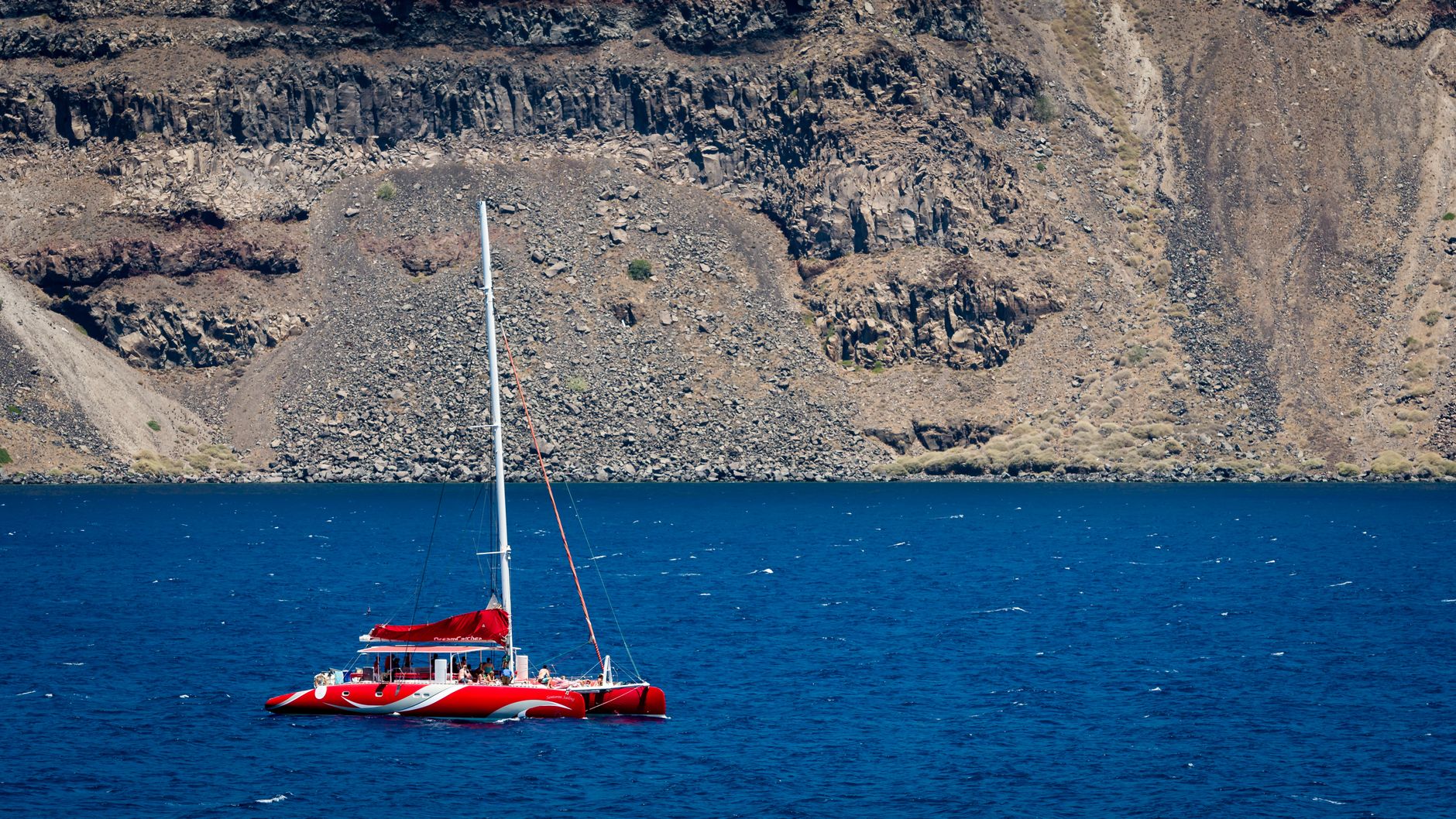 A vibrant red sailboat cruising in the deep blue waters of Santorini with rocky cliffs in the background.